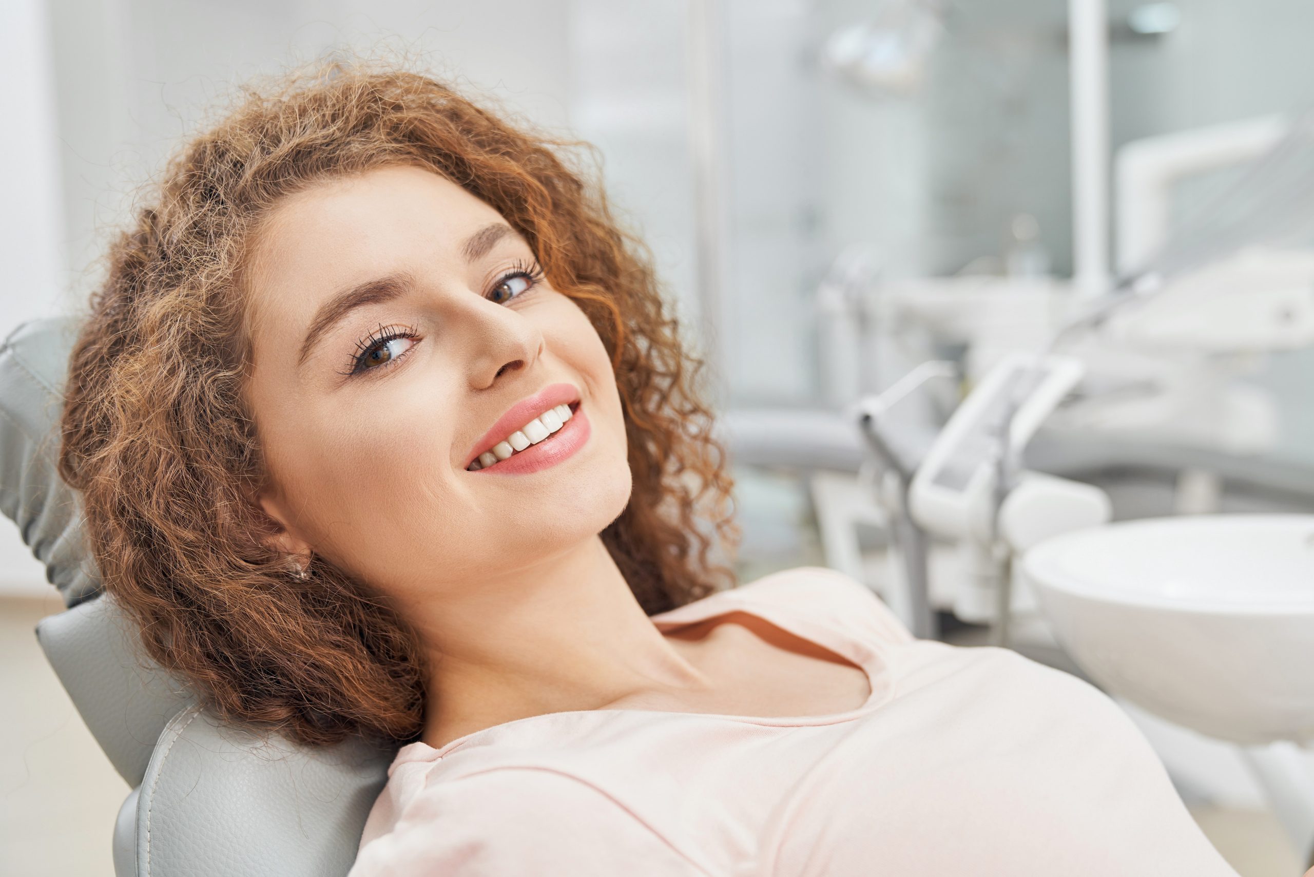 Beautiful female patient with curly hair in pink shirt sitting in dental clinic and waiting for teeth examination, smiling and looking at camera. Concept of medicine and treatment